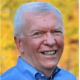 A white man with grey hair wears a blue button-up as he smiles and poses for a photo.